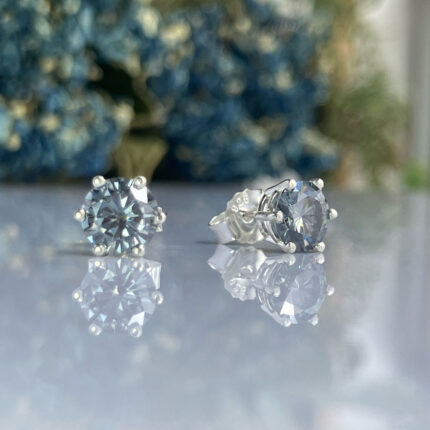 Pair of round-cut diamond stud earrings on a reflective surface with a blurred blue-green background.