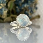 Silver ring with a round milky opal-like gemstone in a beaded bezel, reflected on a glossy surface.