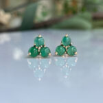 Emerald-green cluster stud earrings with three stones set in gold prongs, displayed on a reflective surface