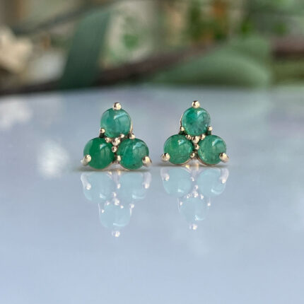 Emerald-green cluster stud earrings with three stones set in gold prongs, displayed on a reflective surface