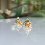 Gold stud earrings in a clover shape with green enamel and turquoise centers, reflected on a glossy surface.