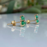 Gold-tone stud earrings with three emerald-green cabochon stones, resting on a reflective surface.
