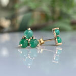 Gold cluster stud earrings with three green cabochon gemstones on a reflective surface.