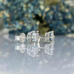 Diamond stud earrings with silver prong settings lying on a reflective surface with a blurred blue background.