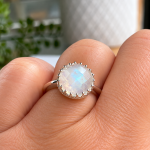 Silver ring with a round iridescent moonstone set in a scalloped bezel on the finger, close-up detail of the gemstone's milky iridescence.