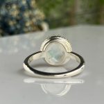Silver ring with oval moonstone bezel-set centerpiece on a glossy surface, showing reflection below.
