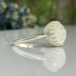 Close-up of a silver ring featuring a faceted round gemstone ball with decorative beaded lace around its circumference on a reflective surface.