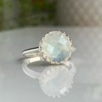 Silver ring featuring a round moonstone cabochon with a faceted surface and a beaded bezel, resting on a reflective surface.