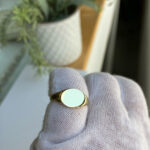 Gold signet ring with a blank white face held on a gloved finger.