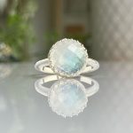 Round faceted moonstone ring in a silver bezel with a beaded edge, reflected on a glossy surface.