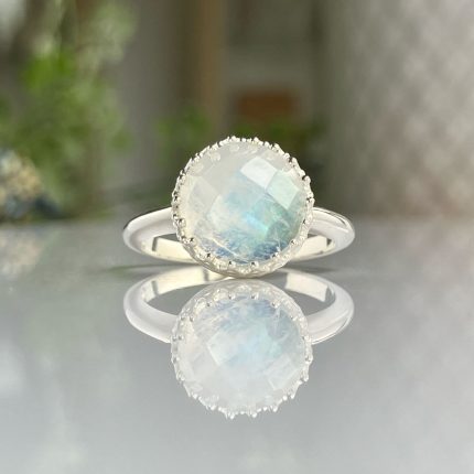 Round faceted moonstone ring in a silver bezel with a beaded edge, reflected on a glossy surface.