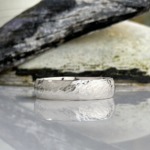Hammered silver wedding band resting on a reflective surface with a rocky backdrop in soft focus.