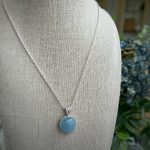 Close-up of a silver chain necklace with a blue pendant resting on beige burlap fabric.