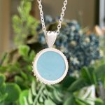 Silver chain necklace with a round blue pendant hanging in focus against a blurred green background