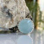 Close-up of a silver chain necklace with a light blue oval pendant set in a beaded bezel, resting against a textured rock surface.