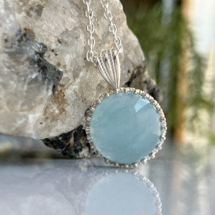 Close-up of a silver chain necklace with a light blue oval pendant set in a beaded bezel, resting against a textured rock surface.