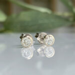 Pair of diamond stud earrings with silver settings resting on a reflective surface, with a blurred green background