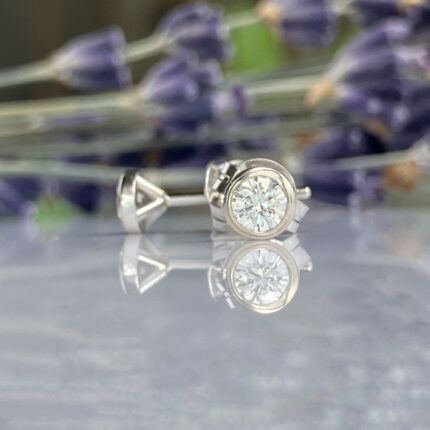 Silver stud earrings with a round-cut diamond on a reflective surface, blurred purple flowers in the background.] , which is invalid formatting