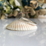 Two silver wedding bands resting on a white ribbed seashell, on a glossy surface with a blurred green background.