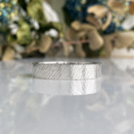 Close-up of a silver textured wedding band resting on a reflective surface with a blurred floral background.