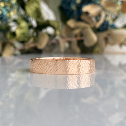 Rose gold textured ring resting on a reflective surface with a blurred floral background, showcasing its hammered finish.