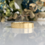 Gold textured band ring resting on a reflective surface with a blurred floral background.