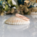 Rose gold hammered wedding band resting on a white seashell with a blurred green background.