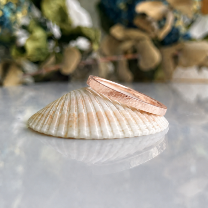 Rose gold hammered wedding band resting on a white seashell with a blurred green background.
