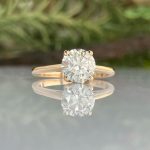 Gold engagement ring with a large round diamond on a polished band, reflected on a glossy surface.