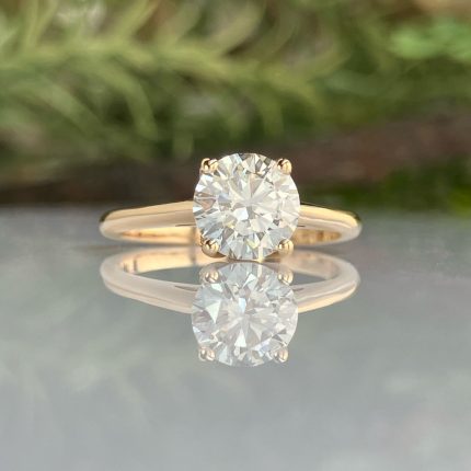 Gold engagement ring with a large round diamond on a polished band, reflected on a glossy surface.