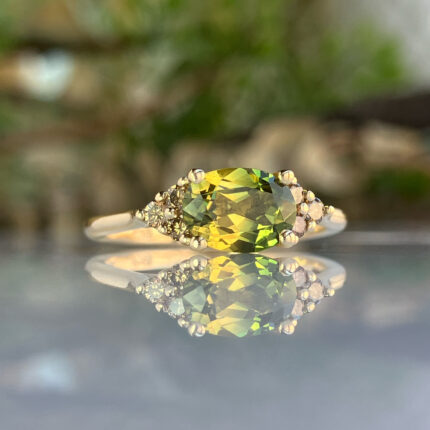 Close-up of a yellow-green faceted gemstone ring surrounded by small crystals, resting on a reflective surface.