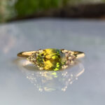 Gold ring with a large green gem surrounded by small diamonds, resting on a reflective surface.