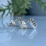 Silver circular stud earrings with an open, lacy edge on a reflective surface with a green plant in the background, in focus mid-shot.