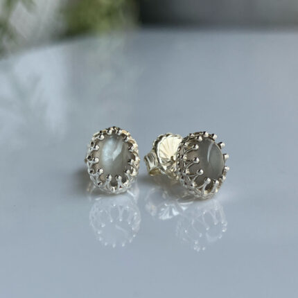 Pair of silver-tone stud earrings with gray cabochon stones in an ornate prong setting, resting on a reflective surface.