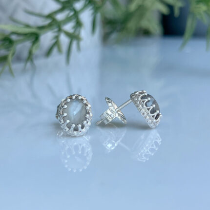 Silver stud earrings featuring a milky moonstone cabochon set in a beaded halo, placed on a reflective surface with greenery in the background.