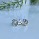 Silver stud earrings with a pale blue cabochon gem set in an intricate beaded bezel, resting on a reflective surface with a butterfly backing visible.