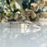 Two silver wedding bands with a brushed finish resting on a reflective surface.