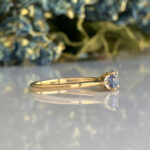 Gold band ring with a small pale blue gemstone in a prong setting on a reflective surface.