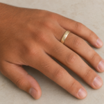 A close-up of a hand wearing a hammered gold wedding band on the ring finger against a beige background.