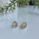 Silver stud earrings with pale gray cabochon stones in a decorative prong frame on a reflective surface.
