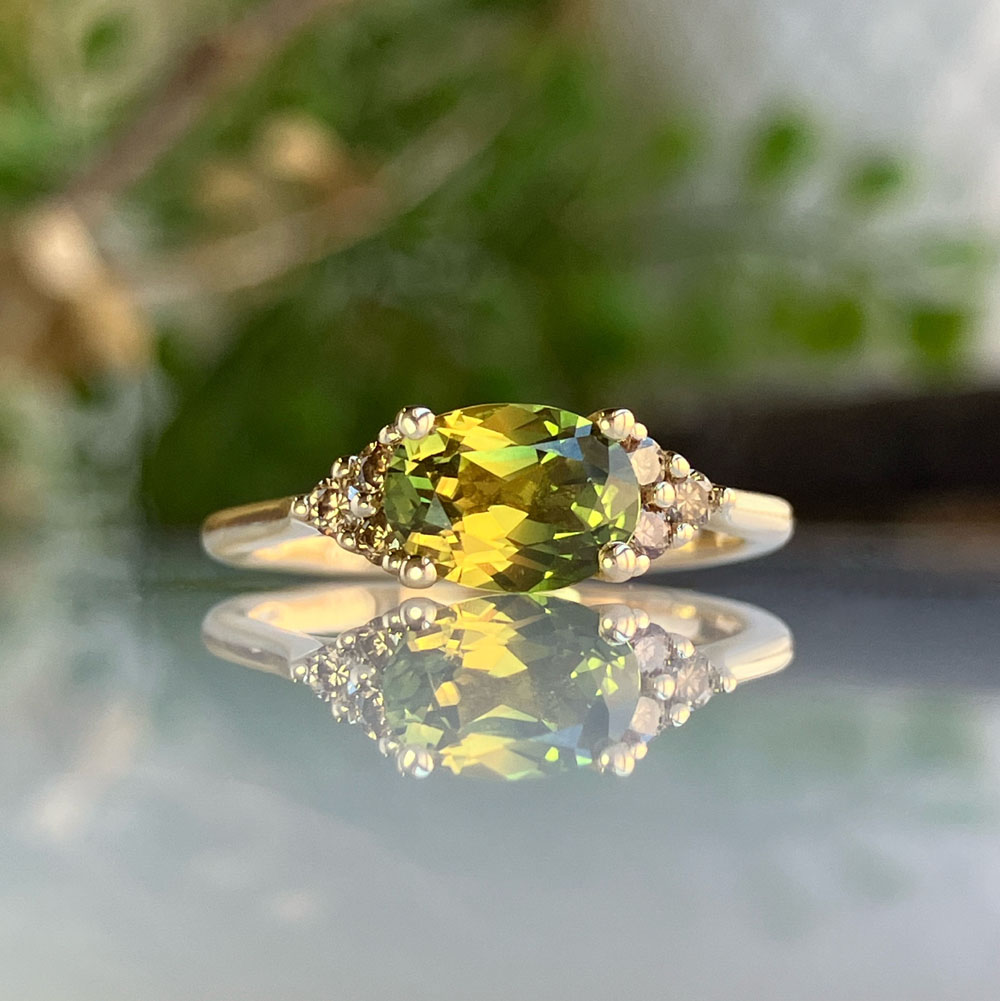 parti australian sapphire engagement ringjpeg | Posy Fine Jewellery Toronto Gold ring with a large oval green-yellow gemstone and small diamonds on the band, reflected on a glossy surface.