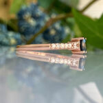 Rose gold ring with a black center gemstone and pave diamonds along the band, reflected on a glass surface.