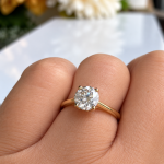 Gold solitaire engagement ring with a round diamond on a finger.
