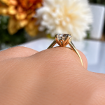 Close-up of a gold engagement ring with a diamond solitaire on a finger, with a blurred floral background.