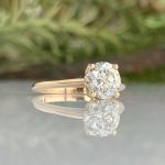 Gold engagement ring with a large round-cut diamond on a reflective surface, greenery blurred in the background.