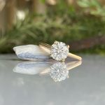 Gold solitaire engagement ring with a round diamond on a yellow-gold band, resting on a pale rock with a soft garden backdrop, and its reflection visible on the surface.