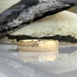 Hammered gold wedding band resting on a reflective surface with a dark, burned-wood background.
