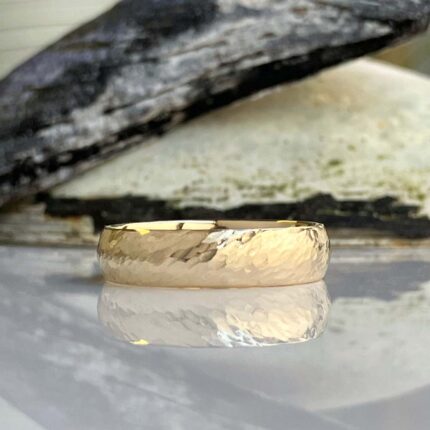 Hammered gold wedding band resting on a reflective surface with a dark, burned-wood background.