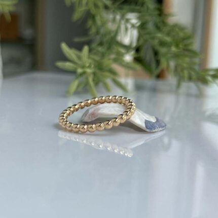 Gold beaded ring resting on a glossy white surface with a small stone and green plant in the background