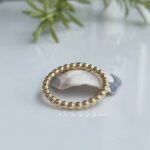 Gold-toned beaded ring resting on a white shell with a soft, blurred green background.